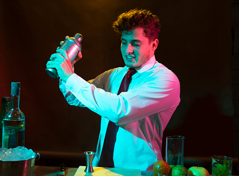 Barman preparing alcoholic drink with a cocktail shaker at the bar with some glasses of liqueur.