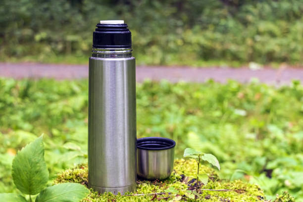 istock Open stainless steel thermos flask with tea drink on stump in coniferous forest. Snack break during hiking concept. 1273021771