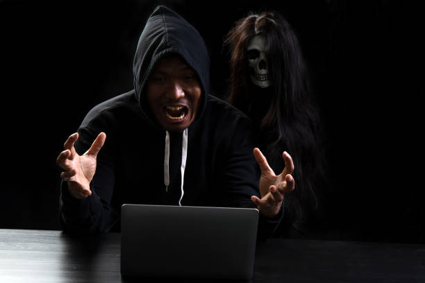 istock Male hacker in hood cloth with skull beside got shocked while using laptop on black background 1273022360