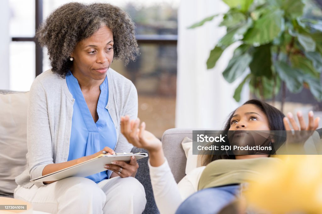 Teen girl gestures from lying down position on therapist's couch The mature adult therapist watches and listens as the teen girl, lying on the couch, gestures as she speaks. Mental Health Professional Stock Photo