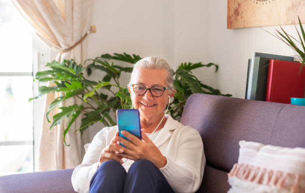 istock Attractive senior lady sitting on sofa at home using her smart phone for chatting with friends or family - woman white hair, glasses wearing earphones - active social and tech people 1273053801