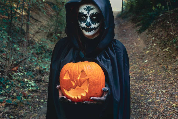검은 후드를 입고 할로윈 페인트 얼굴 마스크를 가진 젊은 여자 - 무서운 마녀 개최 으스스한 갈망 호박 - spookey 뉴스 사진 이미지