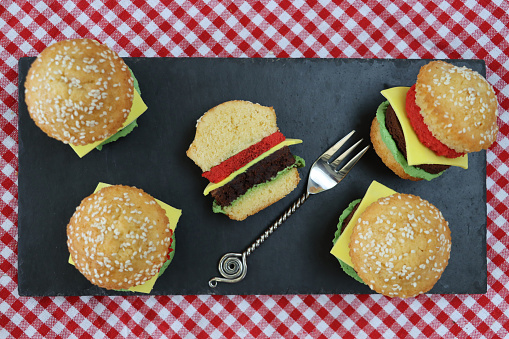 Stock photo showing an elevated view of a group of cupcakes designed as a mini cheeseburgers in a sesame seed buns with chocolate sponge beef burgers, green sponge lettuce, red sponge tomatoes and yellow fondant icing cheese slices. The cakes can be seen in a diner setting on a slate place mat with red and white checked table cloth.