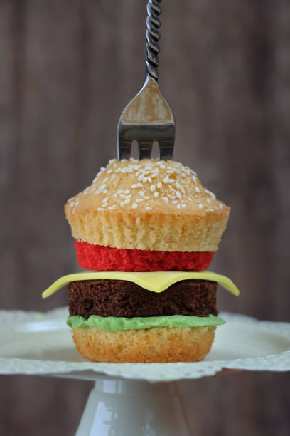 Stock photo showing a close-up view of cupcake designed as a mini cheeseburger in a sesame seed bun with chocolate sponge beef burger, green sponge lettuce, red sponge tomato and yellow fondant icing cheese slice. The cake can be seen with a metal fork protruding from it, in a diner setting on a white cake stand. imagen de pastel, cupcakes caseros de chocolate y vainilla diseñados como una mini hamburguesa con queso en bollo de semillas de sésamo, lechuga esponja y tomate con rodaja de queso glaseado fondant, tenedor de metal, fondo marrón, enfoque en primer pla