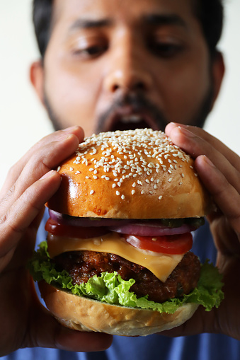 Stock photo showing close-up of asian man about to eat a homemade beef burger served in a sesame seed bread bun with melted cheddar cheese slice, shredded lettuce, tomatoes and gherkin slices and rings of red onion.