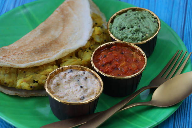 Stock photo showing a close-up view of Indian dosa pancakes on a green, banana leaf design plate with gold cutlery and dishes of tasty sambar, coconut chutney, mint and coriander chutney, ginger, chilli and garlic dip, and peanut chutney served. Savoury crepes with masala potato filling, against a blue wood grain background, popular in India / Pakistan. imagen de verde, placa de diseño de hoja de plátano de tortitas saladas de dosa india, crepe de panqueque relleno con puré de patata masala, cuencos de chutney de coco, chutney de menta y cilantro, jengibre, chile y ajo dip, y chutney de cacahuete, fond