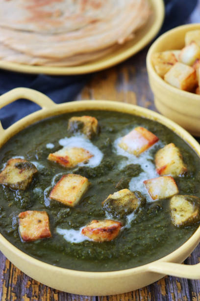 Stock photo showing close-up view of Palak paneer being served in a yellow serving dish with handles. This is an Indian vegetarian dish consisting of paneer, similar to cottage cheese, in pureed spinach flavoured with spices including ginger, garlic and garam masala. imagen de primer plano de mango, plato amarillo plato vegetariano comida vegetariana palak paneer (queso de algodón y puré de espinacas) receta junto a tela de queso azul-gris, lachha paratha (pan plano) y tazón de queso de cuajada en cubos, fondo de me