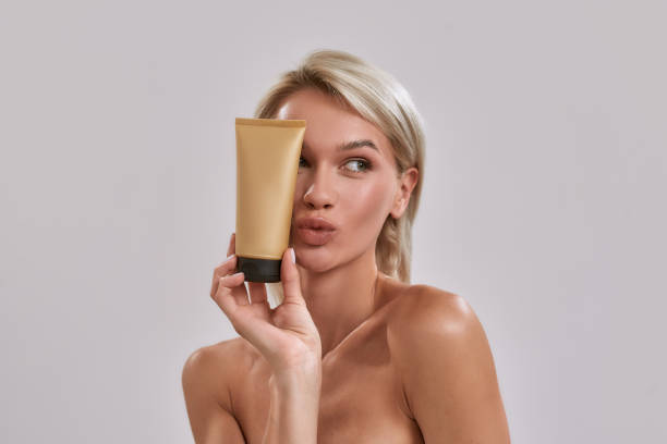 Portrait of cheerful young woman with perfect glowing skin looking aside, holding sunscreen skincare product while posing isolated over grey background Portrait of cheerful young woman with perfect glowing skin looking aside, holding sunscreen skincare product while posing isolated over grey background, Horizontal shot tanned body stock pictures, royalty-free photos & images