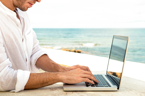 Detail of hands of guy working on the notebook digital device at the seaside wearing white shirt sunny day