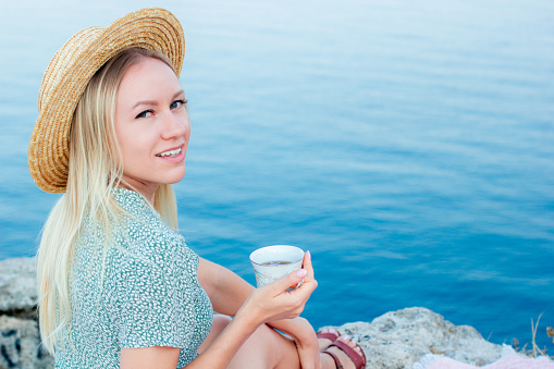 A woman with blond hair sits on the seashore with a glass in her hands.