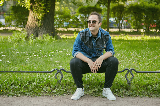 Smiling young man in sunglasses is sitting on the fence in the public park. Male tourist has a rest outdoors.