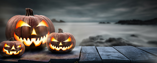 Three spooky halloween pumpkins, Jack O Lantern, with an evil face and eyes on a wooden bench, table with a misty gray coastal night background with space for product placement.