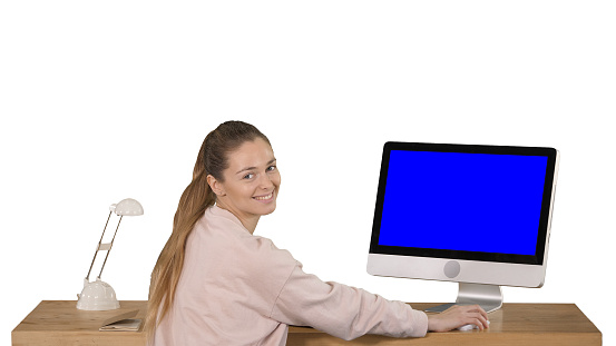 View to the screen of computer. Cheerful woman sitting at the table with a computer in office and looking at camera smiling Blue Screen Mock-up Display on white background. Professional shot in 4K resolution. 010. You can use it e.g. in your commercial video, business, presentation, broadcast