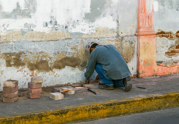 Mazatlan, Mexico - April 23, 2008: Closeup of kneeled male worker, dressed in blue, repairing gray pavement of sidewalk downtown. Redish stones, white wall, greenish mortar.