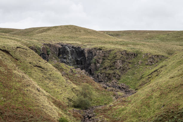 Wild waterfall flowing over rocks and grassy hills surrounding Wild view of distant waterfall at Glenariff forest in County Antrim coast, Northern Ireland, in summer with grassy hills and a cloudy sky glenariff photos stock pictures, royalty-free photos & images