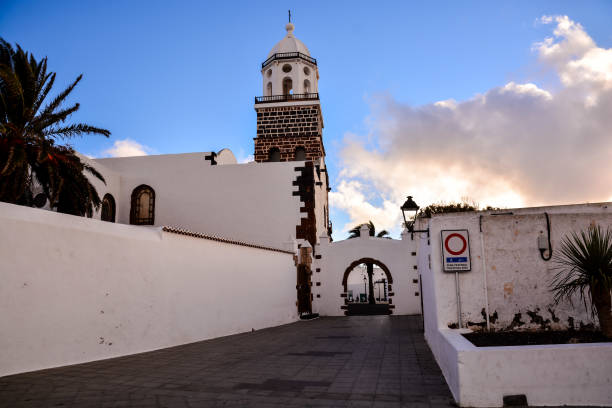 Teguise, Lanzarote, Canary Islands, Spain - March 4th 2025: Two travelers stand at a Spanish bus stop, closely examining the posted schedule as they plan their next destination. The setting includes a white wall with a green door, creating a classic Mediterranean ambiance. The cloudy sky adds a subdued atmosphere, while the paved road and urban infrastructure highlight the practical side of exploring a foreign city. Their backpacks and casual attire reinforce the theme of independent travel, capturing the essence of adventure and discovery.