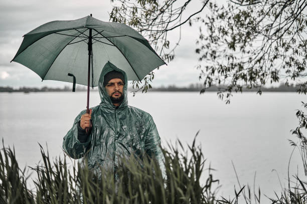 a man in a green raincoat and with an umbrella on the shore near the water. mainly cloudy - 11275 imagens e fotografias de stock