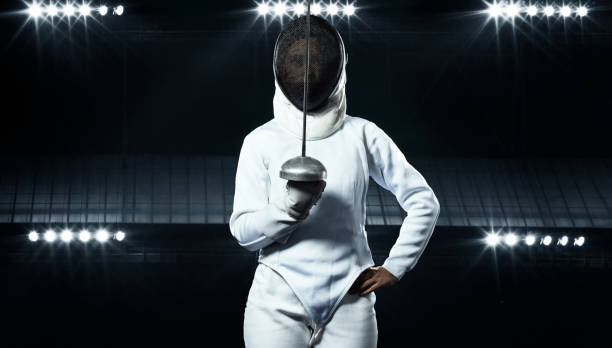 Young fencer athlete wearing fencing costume holding the sword and mask. Black background with lights stock photo