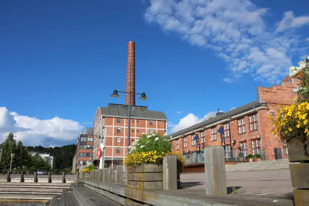 Photo of Wooden embankment with steps and flower beds against the background of a red brick factory with a large chimney on the shore of lake Vesiyarvi in the city of Lahti. Finland.