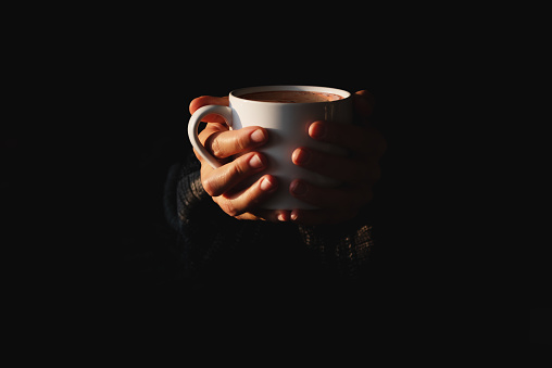 Female hands holding white cup of coffee on black background. Woman warming her hands on mug of hot beverage in the dark. Copy space. Minimal style. Low key