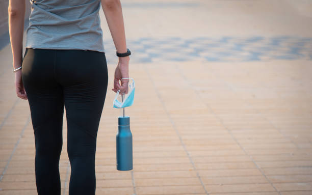 Woman carrying water bottle and protective surgical face mask before jogging on the running track for preventing coronavirus spread Woman carrying water bottle and protective surgical face mask before jogging on the running track for preventing coronavirus spread Covid-19 pandemic spread do a safe exercise routine yoga pants stock pictures, royalty-free photos & images