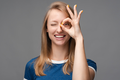 Cheerful smiling female with blonde straight hair, demonstrating white teeth, looking at the camera through fingers in OKAY gesture. Face expressions, emotions concept.