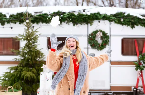 Happy Cheerful Young Woman Throwing Snowball At Camera, Playing Snow Fight With Somebody, Enjoying Camping Vacation In Winter Campsite, Having Fun Outdoors In Woods With Modern Trailer On Background