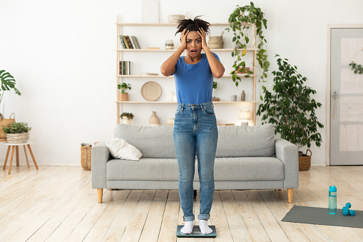 African American Woman Stressing Out About Gaining Excess Weight Standing On Scales Posing At Home, Looking At Camera. Empty Space