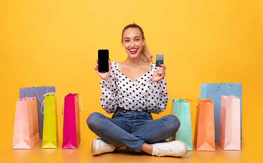 Online Shopping Concept. Excited overjoyed lady showing smartphone and credit card to camera, ordering clothes and purchases via internet, sitting with colorful paper bags at pastel orange studio