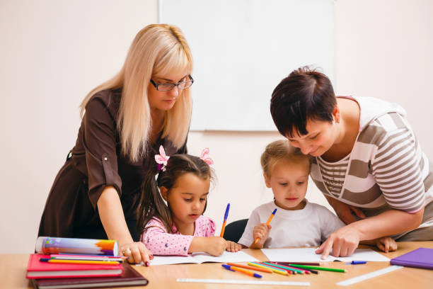istock Two teachers in class with their young students with a beautiful young woman helping two small girls at a desk 1273276217