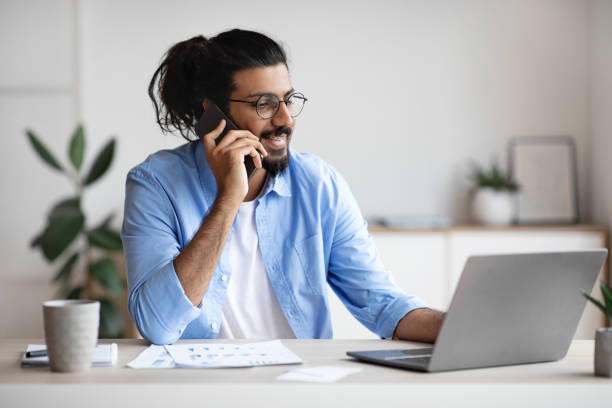 istock Business Communication. Young Indian Male Entrepreneur Talking On Cellphone While Working On Laptop 1273276232