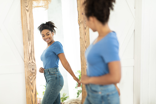 Perfect Weight. Cheerful Slim Black Girl After Slimming Looking At Reflection In Mirror Standing Indoor. Selective Focus