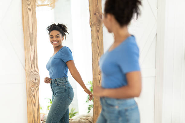 fröhliche afroamerikanische mädchen nach abnehmen blick in spiegel indoor - abnehmen fotos stock-fotos und bilder