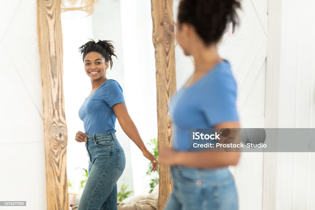 Joyful African American Girl After Slimming Looking In Mirror Indoor Successful Weight Loss. Joyful African American Girl After Slimming Smiling To Her Reflection In Mirror Standing At Home. Staying Fit, Dieting And Weight-Loss Concept. Selective Focus Dieting Stock Photo