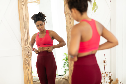 Infeliz mujer africana tocando el vientre gordo de pie cerca de espejo interior photo
