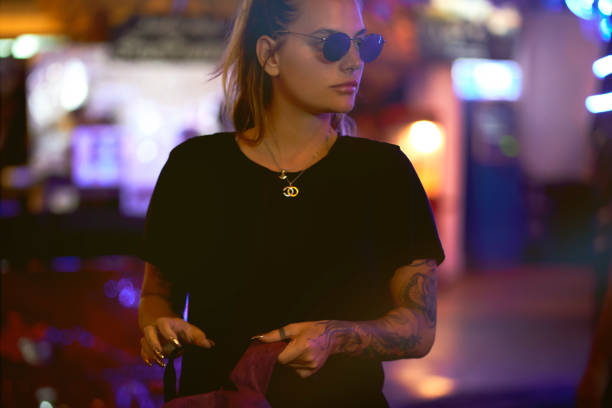 Cool young girl with black t-shirt lit by neon lights at night. Cool young girl with tattoos and sunglasses and black t-shirt lit by neon lights at night. black tattoo stock pictures, royalty-free photos & images