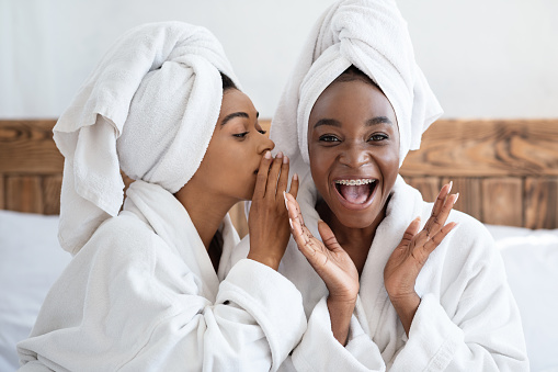 Two pretty african american young women having girls day at home, wearing bathrobes, sitting on bed. Black woman whispering secrets to her emotional bestie, bedroom interior, closeup portrait