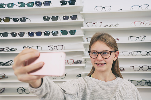 Nice girl with new eyeglasses. Holding her smartphone and smiling to the camera. Fashionable children's eyeglasses. Video conference with her mother to confirm the choice.