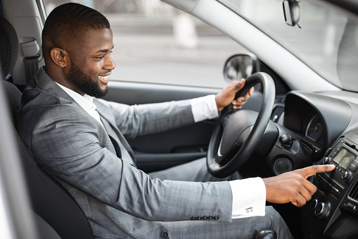 Smiling black businessman in suit turning on music in car before moving. Cheerful african american guy driver pushing button on functional panel in his car, turning on music before moving, side view