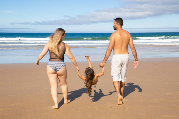 Parents and little kid wearing swimsuits Parents and little kid wearing swimsuits, walking on golden sand to water. Girl holding parents hands, hanging and throwing legs up. Rear view. Family outdoor activities concept backside for a girl on the beach stock pictures, royalty-free photos & images