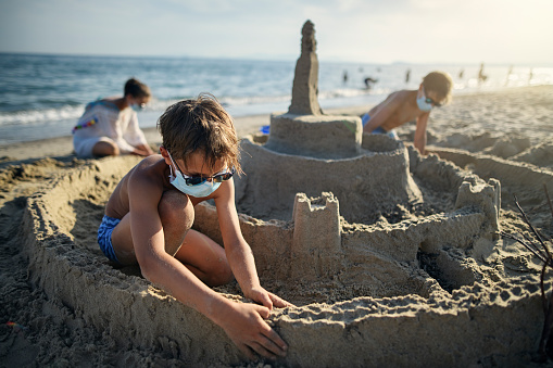 Three kids are having fun building a huge sandcastle on a beach on a sunny summer day. \nCOVID-19 pandemic and the kid are wearing surgical masks.\nNikon D850