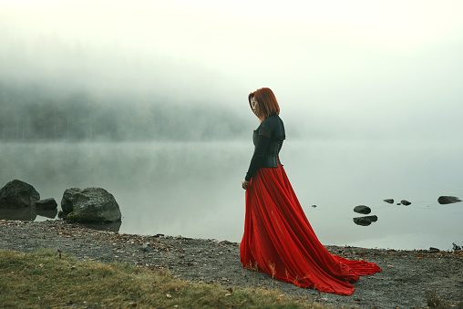 Redhead posing in red dress at the lake with fog, feeling lonely and sad