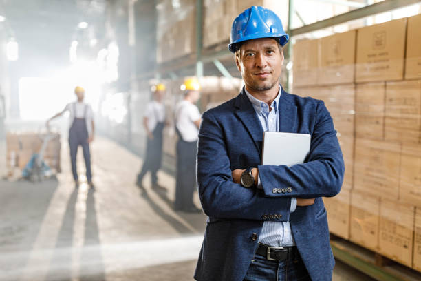 mid adult foreman with crossed arms in a warehouse. - blue helmets imagens e fotografias de stock