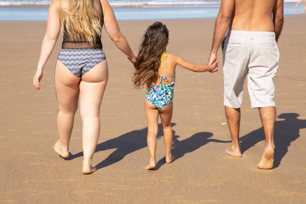 Parents and kid wearing swimsuits, walking to water Parents and kid wearing swimsuits, walking to water on golden sand. Girl holding parents hands. Rear view. Family outdoor activities concept backside for a girl on the beach stock pictures, royalty-free photos & images