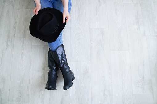 Woman legs in black cowboy boots with a cowboy hat. Sitting on a gray wooden background.