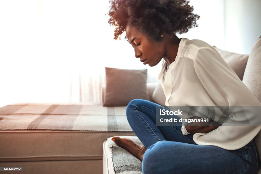 Woman sitting on bed and having stomach ache. Sad beautiful woman sitting on the couch and feeling spasm and symptoms of pms. Shot of a young woman experiencing stomach pain on the sofa at home Pain Stock Photo