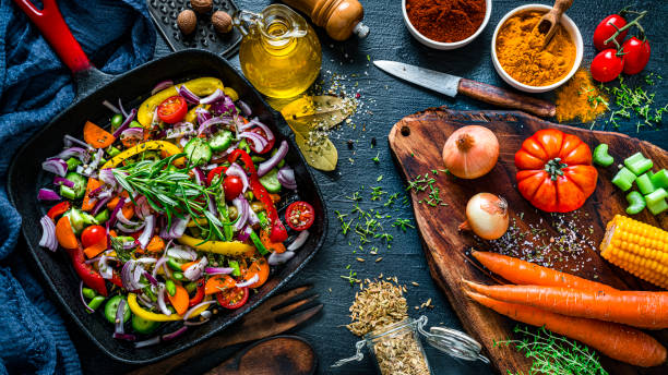 Healthy food backgrounds: overhead view of a rustic dark kitchen table filled with healthy fresh organic vegetables and spices. A cast iron cooking pan is at the left with sliced vegetables. A cutting board is at the right. Vegetables and spices included in the composition are bell pepper, tomatoes, celery, broccoli, onion, carrots, corn, rosemary, turmeric, paprika, dried parsley, nutmeg, peppercorns, salt, bay leaves, among others. High resolution 42Mp studio digital capture taken with Sony A7rII and Sony FE 90mm f2.8 macro G OSS lens