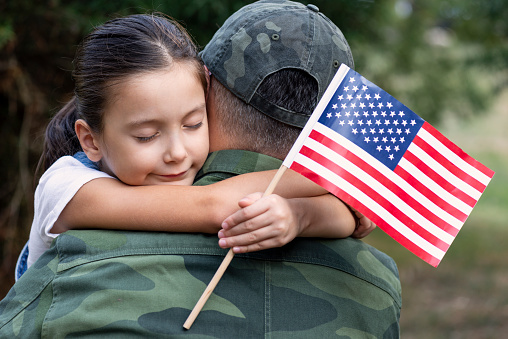 Little girl hugs the soldier father and holding American flag.