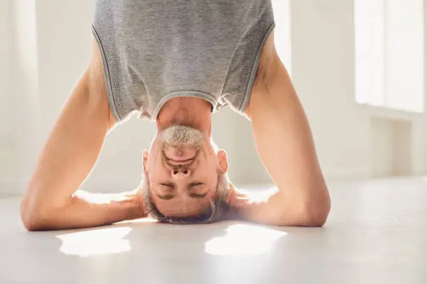 Mature gray haired male in sportswear doing salamba sirsasana or supported headstand exercise while practicing yoga in light room