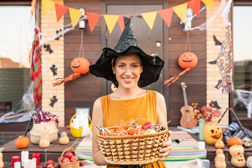 Young pretty woman in black hat of halloween witch holding basket with sweets while waiting for halloween kids against decorated door
